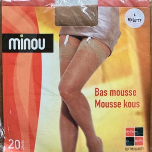 Puede incluir: Paquete de medias transparentes de color marrón claro. La marca "minou" aparece en letras blancas sobre un rectángulo negro. El paquete también presenta las palabras "Bas mousse" y "Mousse kous". Las medias son talla L.