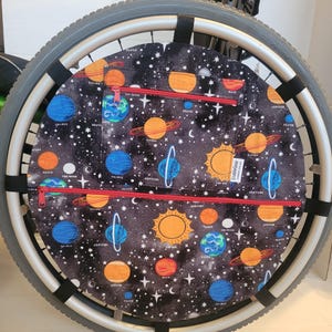 Puede incluir: Cubierta de rueda de silla de ruedas con diseño espacial. La cubierta presenta planetas, estrellas y el sol sobre un fondo negro. Tiene dos bolsillos rojos con cremallera y se sujeta a la rueda con correas negras.