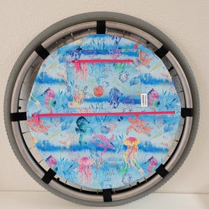 May include: A wheelchair wheel cover with a vibrant aquatic design. The cover features a blue background with colorful illustrations of sea turtles, jellyfish, and coral. It has two zippered pockets with pink zippers and black straps for attachment.