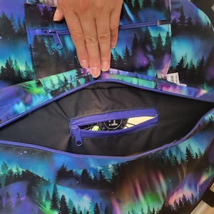 May include: A large, round bag with a vibrant, colorful design featuring a forest silhouette against a backdrop of aurora borealis. The bag has multiple zippered compartments, with a blue zipper and a black interior.