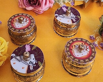May include: Four small round gold boxes with ornate designs. Each box has a white cow with a floral design on the lid. The boxes are on a yellow textured surface.