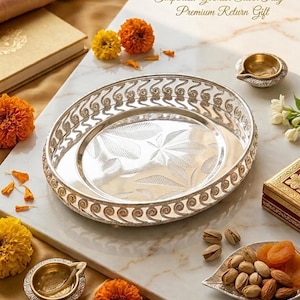 May include: Oval German silver tray with intricate border design and leaf pattern. The tray is surrounded by orange and yellow flowers, small brass oil lamps, and a leaf-shaped dish filled with nuts and dried apricots. The text on the tray says "Imported German Silver Tray - Premium Return Gift."
