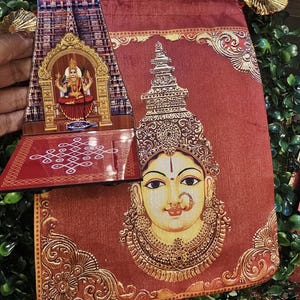 May include: A rectangular fabric pouch in a deep red hue featuring a printed illustration of a deity's face with ornate gold jewelry. Beside it, a flat acrylic stand displays a detailed image of a temple tower and a seated deity with a white kolam.