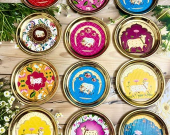 May include: A collection of decorative round trays with gold rims. Each tray features a unique colorful design, including floral patterns and illustrations of a cow. The trays are arranged on a wooden surface, showcasing their vibrant colors and intricate details.