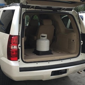 May include: A white SUV with its rear hatch open, revealing the interior. A large, white propane tank sits on a black base in the cargo area. The vehicle's interior features tan seats and trim.
