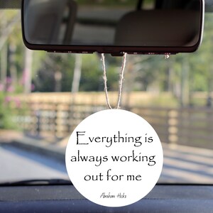 May include: White round car air freshener with the text "Everything is always working out for me" and the name "Abraham Hicks" at the bottom.