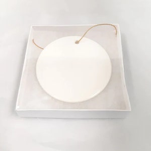 May include: A white ceramic ornament with a gold string attached. The ornament is round and has a smooth surface.