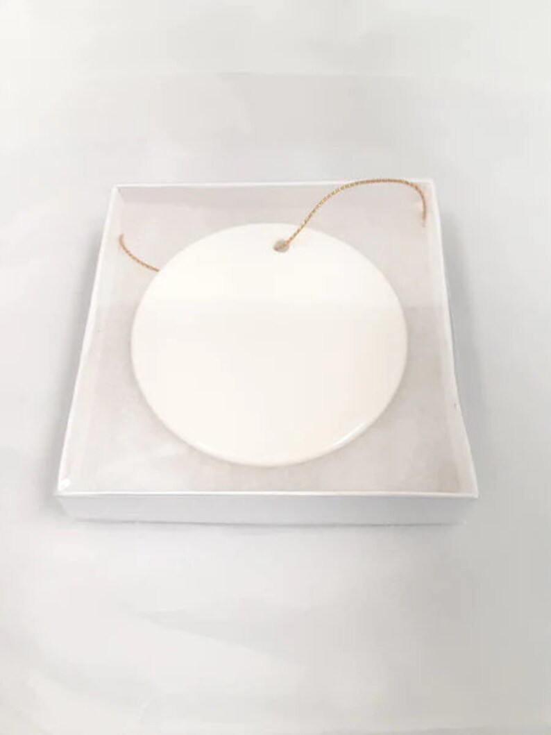 May include: A white ceramic ornament with a gold string attached. The ornament is round and has a smooth surface.