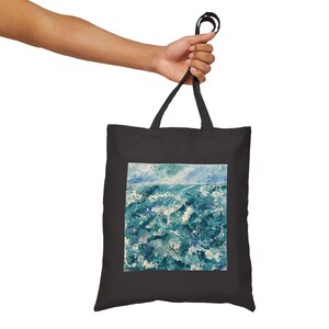 Puede incluir: Bolso tote negro con una pintura de un paisaje marino. La obra de arte representa un océano agitado en tonos azules y blancos, bajo un cielo azul claro. El bolso es sostenido por una mano.
