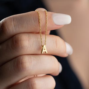 May include: A gold-colored necklace with a pendant in the shape of the letter "A". The chain is delicate, and the pendant is small and centered. The necklace is held up by a hand with manicured nails.