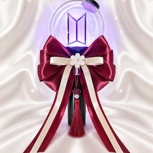 May include: A clear, spherical light stick with a purple glow and a stylized letter design. It is adorned with a large burgundy bow, cream-colored ribbons, and a tassel. The light stick is set against a white, silky background.