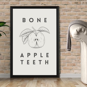 May include: Black framed art print featuring a detailed illustration of a sliced apple with leaves. The text "BONE APPLE TEETH" is printed above and below the apple illustration. The artwork is displayed against a light background.