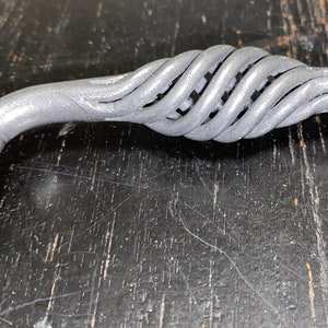 Puede incluir: Un tirador de armario de metal plateado con un diseño retorcido en forma de cuerda. El tirador tiene un acabado liso y pulido.