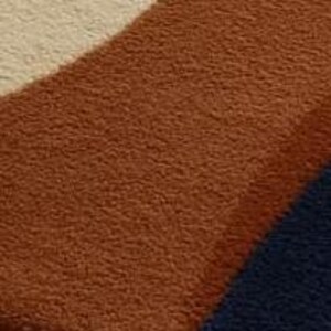 Puede incluir: Primer plano de una alfombra suave y texturizada con un dise&ntilde;o geom&eacute;trico. La alfombra presenta una secci&oacute;n curva en un color marr&oacute;n rojizo c&aacute;lido, una secci&oacute;n de color crema y una secci&oacute;n triangular azul oscuro. La alfombra parece afelpada y acogedora.