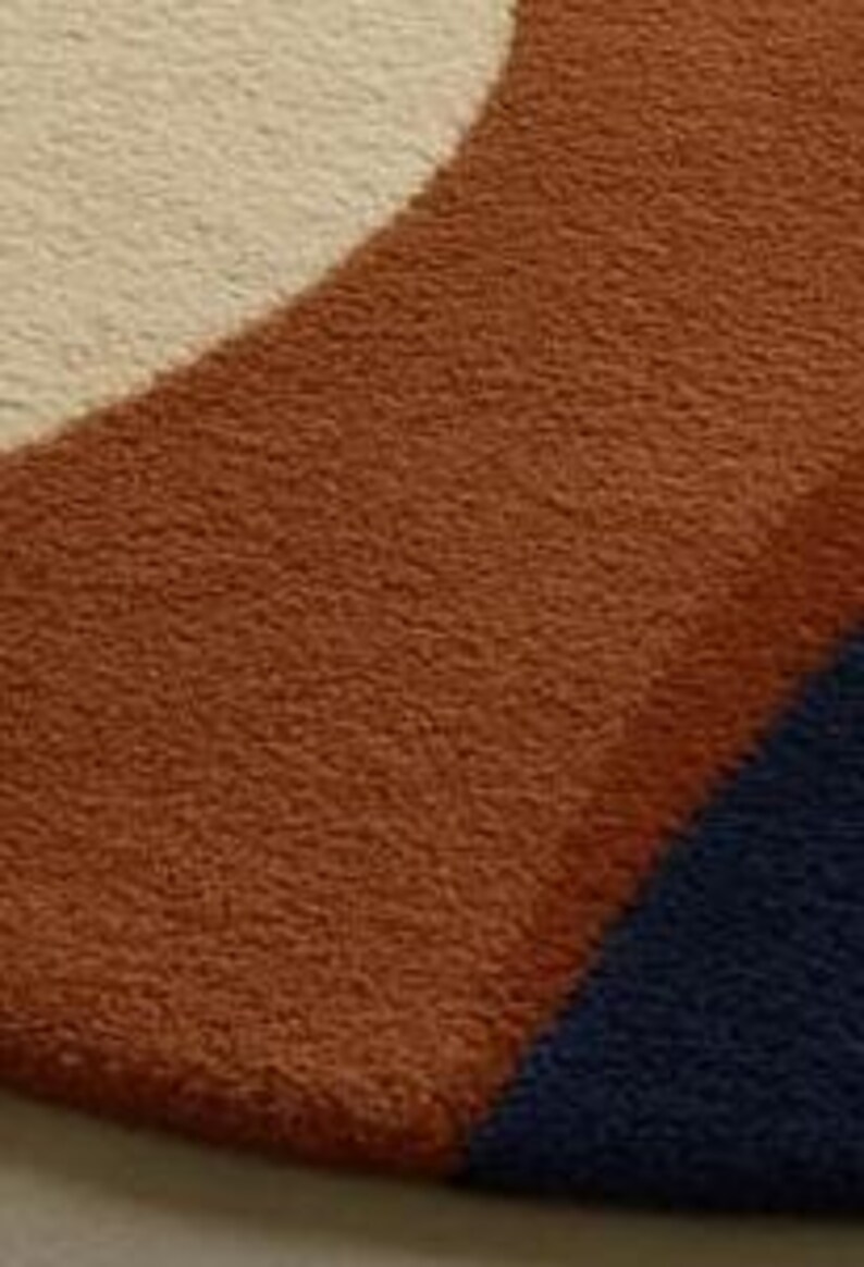 Puede incluir: Primer plano de una alfombra suave y texturizada con un dise&ntilde;o geom&eacute;trico. La alfombra presenta una secci&oacute;n curva en un color marr&oacute;n rojizo c&aacute;lido, una secci&oacute;n de color crema y una secci&oacute;n triangular azul oscuro. La alfombra parece afelpada y acogedora.