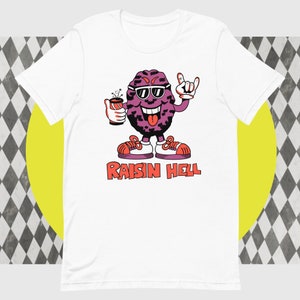 Puede incluir: Camiseta blanca con una uva de dibujos animados que lleva gafas de sol y sostiene una lata de refresco. La uva está haciendo un gesto de rock and roll. El texto "RAISIN HELL" está impreso debajo de la uva.
