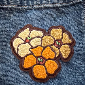 May include: A textured, three-flower embroidered patch featuring cream, golden-yellow, and orange petals with dark brown outlines. The floral design is stitched onto a blue denim fabric background, showcasing a dense, looped thread construction.