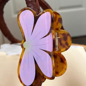 May include: A flower-shaped hair clip with a tortoiseshell pattern and a lavender center. The clip is made of a glossy material and has a metal spring mechanism. The hair accessory is attached to a brown leather handle.