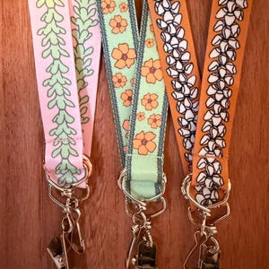 May include: Close-up of three lanyards with different patterns. One is pink with green leaves, another is light green with orange flowers, and the last is orange with white floral designs. Each lanyard has a metal clip and ring.