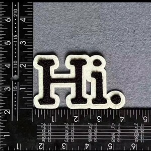 May include: A black and white patch with the word "Hi" in a bold, block letter style. The letters are black with a white border. The patch is on a gray surface.