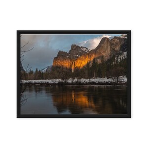 May include: A scenic view of a waterfall cascading down a rocky cliff face in Yosemite National Park. The water is illuminated by the setting sun, creating a warm glow. The river in the foreground reflects the golden light, and the surrounding mountains are covered in snow.
