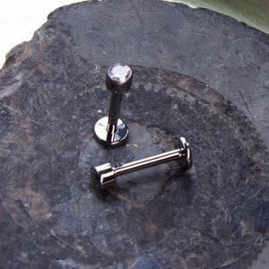 May include: Two silver body jewelry pieces, one with a clear gem and the other a plain bar, on a dark gray stone surface.