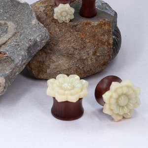 May include: Three pairs of wooden ear gauges with white flower designs. The gauges are displayed on a rock.