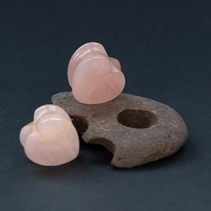 May include: Three pink heart-shaped rose quartz stones on a gray rock. The stones are smooth and polished.