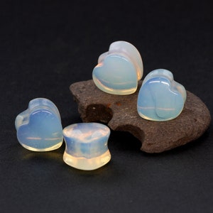May include: Four heart-shaped opal plugs, two are on a dark gray stone, and two are on a black surface.