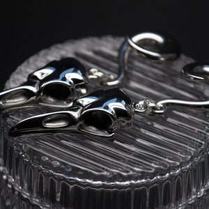 May include: A pair of silver earrings featuring a detailed raven skull design. The earrings are made of a smooth, polished metal and have a unique, curved shape.