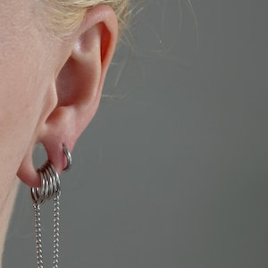 Puede incluir: Primer plano de una oreja adornada con pendientes plateados. El diseño incluye múltiples aros apilados y una cadena. También es visible un pequeño pendiente de aro. El estilo es minimalista y moderno.