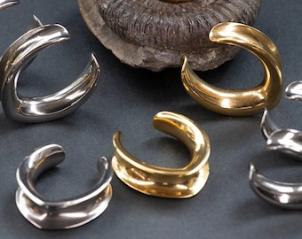 Puede incluir: Una colección de dilatadores de oreja en forma de media luna en tonos oro y plata pulidos. Estas piezas de joyería corporal curvas y lisas presentan un diseño abierto, mostrando un acabado metálico que refleja la luz en sus superficies redondeadas.