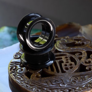 Peut inclure: Deux écarteurs d'oreille noirs empilés sur une surface décorative sculptée. Les écarteurs ont une finition lisse et brillante et un design circulaire. L'arrière-plan présente une vue floue d'autres objets, ajoutant de la profondeur à l'image.