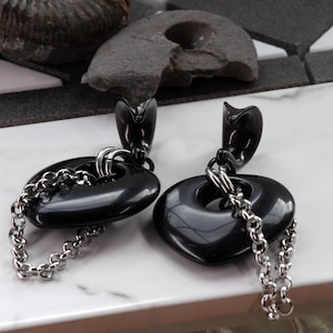 May include: A pair of black oval earrings with a silver chain. The earrings have a glossy finish and a unique design. The chain is attached to the oval shape and hangs down. The earrings are displayed on a white surface.