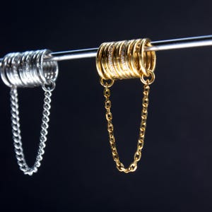 Pode incluir: Close-up de dois conjuntos de joias. Um conjunto é prateado, com uma corrente e vários anéis. O outro conjunto é dourado, com uma corrente e anéis, alguns incrustados com pequenas pedras transparentes. Ambos os conjuntos estão pendurados numa barra de metal contra um fundo escuro.