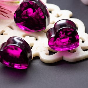 May include: Three heart-shaped, translucent, fuchsia-coloured objects with dark interior swirls. They rest on a white, carved, decorative surface. A pink flower is visible in the background. The objects appear to be made of resin or glass.