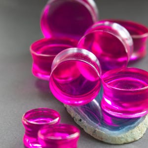 May include: A collection of translucent, vibrant hot pink ear plugs with a smooth, cylindrical shape and flared edges. These circular body jewelry pieces are arranged in a cluster, showcasing their bright magenta color and glossy finish.