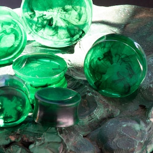 May include: A selection of green ear plugs with a marbled design. The plugs are in various shapes and sizes, with a shiny finish. The plugs are displayed on a textured, green surface.