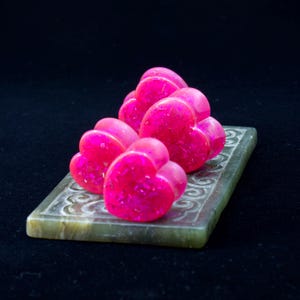May include: Two bright neon pink heart-shaped ear plugs with embedded iridescent glitter flakes. The heart-shaped jewellery pieces are arranged in a pair, showcasing a glossy finish and a vibrant, saturated magenta colour throughout the material.