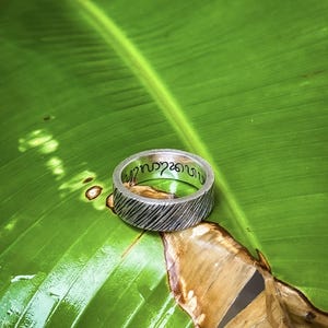 May include: A silver ring with a textured band and an inscription in Thai script. The ring is resting on a green leaf.