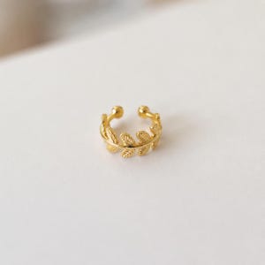 Pode incluir: Um ear cuff dourado com um padrão delicado e repetitivo de folhas ao longo da banda. A peça de joalharia tem um design aberto com pequenos terminais arredondados em cada extremidade e um acabamento metálico texturizado em toda a estrutura.