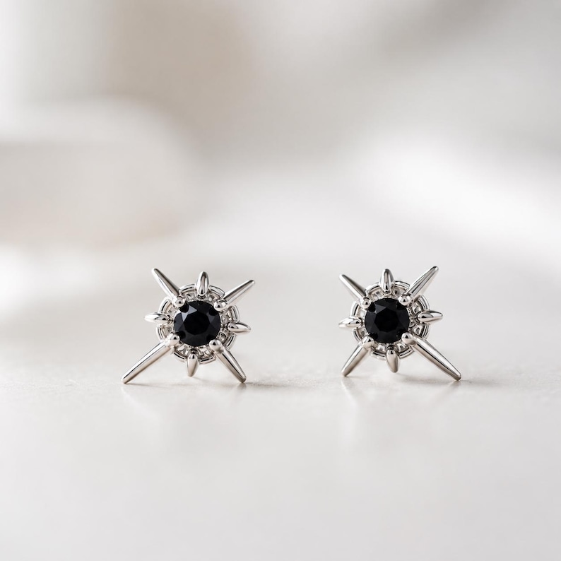 Puede incluir: Un par de pendientes de bot&oacute;n de plata con una piedra negra redonda central rodeada por un dise&ntilde;o de estrella con m&uacute;ltiples puntas radiantes. El engaste de metal es de plata pulida y los pendientes tienen una forma de estrella sim&eacute;trica.