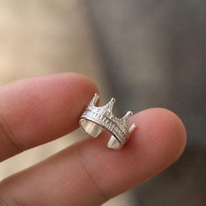 Könnte beinhalten: Ein silberfarbener Ohrring in Kronenform mit einer texturierten Oberfläche und kleinen, reflektierenden Details entlang des Bandes und der Spitzen. Das Schmuckstück hat ein offenes Design, wodurch es individuell am Ohrknorpel angepasst werden kann.