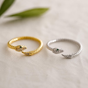 May include: Two open-style rings shaped like snakes, one in gold tone and one in silver tone. Each ring features a textured scale pattern, small clear stones along the head, and a single tiny green stone representing the eye of the snake.