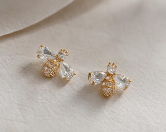 May include: A pair of gold-toned bee-shaped stud earrings featuring clear, pear-shaped and round-cut crystals. The body of each bee is encrusted with small, sparkling white stones, while the wings are formed by larger, faceted teardrop crystals.