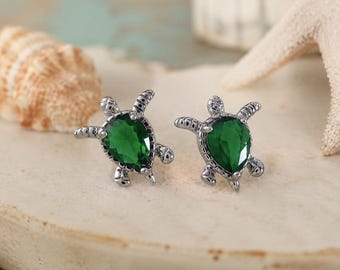 May include: A pair of silver-toned stud earrings shaped like sea turtles, each featuring a pear-shaped, deep green faceted gemstone set into the shell area. The metal has a textured, dark silver finish detailing the head, flippers, and tail.