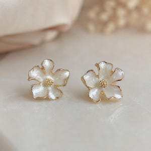 May include: A pair of stud earrings shaped like five-petaled flowers. Each earring features white, pearlescent enamel petals with irregular, wavy edges and a gold-toned metal outline. The center of each flower has a small, textured gold-toned stamen.