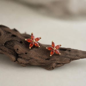 May include: A pair of stud earrings shaped like five-petaled flowers. Each earring features five marquise-cut orange gemstones arranged around a central gold-toned metal bead, creating a floral design with a bright, warm colour palette.