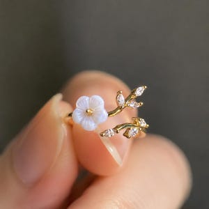 Può includere: Anello dorato con un delicato design floreale. L'anello presenta un fiore bianco e un ramo con foglie e piccole pietre trasparenti. L'anello è tenuto da una persona. Gioiello elegante.