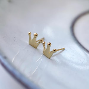 Könnte beinhalten: Ein Paar goldene Kronen-Ohrstecker. Die Ohrringe sind klein und zart und wären ein tolles Geschenk für einen besonderen Anlass.